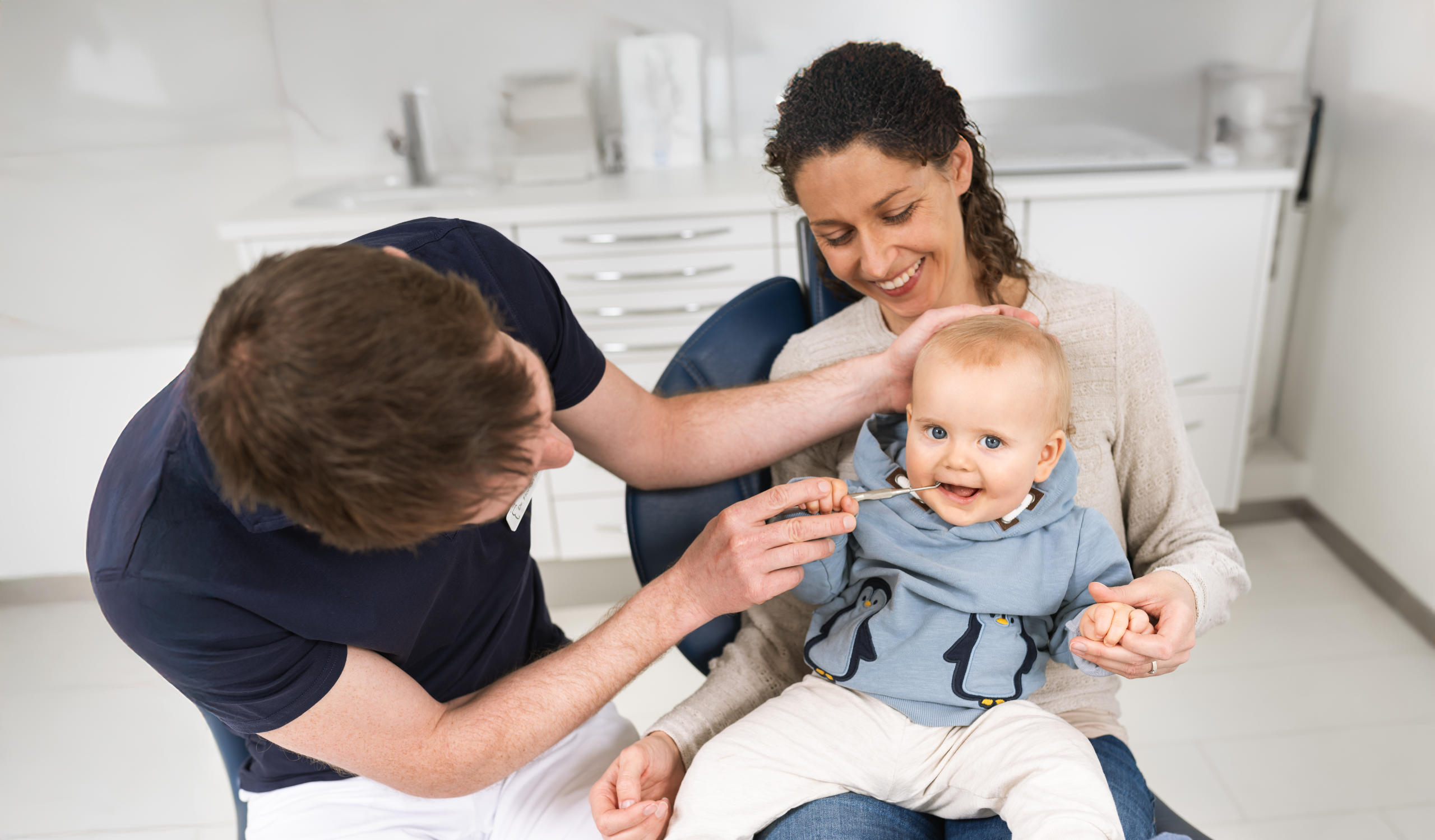Eine Mutter mit dunklen Haaren sitzt mit ihrem Baby auf dem Behandlungsstuhl. Dr. Steib sitzt daneben und untersucht das lächelnde Baby mit Zahnarztbesteck