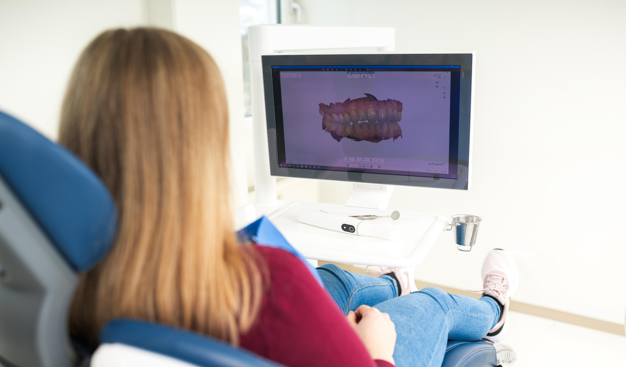 Ansicht von Hinten auf eine Frau in einem roten Pullover, welche auf dem Behandlungsstuhl sitzt und auf ein Display schaut, auf welchem ein 3D Scann farbig abgebildet ist. Darunter liegt ein weißer Intraoralscanner auf einem weißen Tisch
