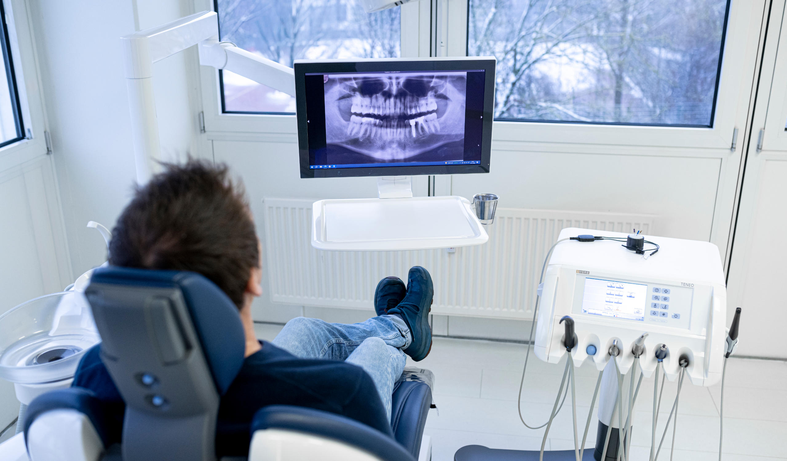 Ansicht von Hinten auf einen Patienten, welche auf dem Behandlungsstuhl sitzt und auf ein Display schaut, auf welchem ein Röntgenbild eines Kiefers abgebildet ist